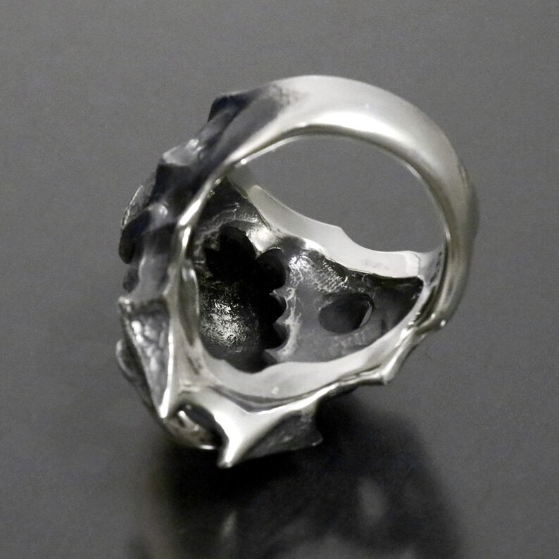 May include: A silver ring with a detailed, textured design. The ring is open on the back, revealing a hollow interior.
