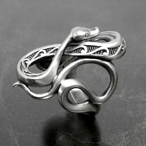 Op de afbeelding: Een zilveren ring met een gedetailleerd slangendesign. De slang is om zichzelf gewikkeld, met zijn kop omhoog en zijn staart eronder gekruld. De ring is ingewikkeld gemaakt met een getextureerd oppervlak en ingewikkelde details.