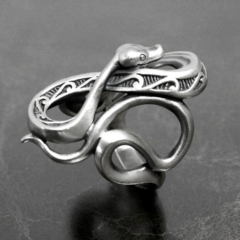 Op de afbeelding: Een zilveren ring met een gedetailleerd slangendesign. De slang is om zichzelf gewikkeld, met zijn kop omhoog en zijn staart eronder gekruld. De ring is ingewikkeld gemaakt met een getextureerd oppervlak en ingewikkelde details.
