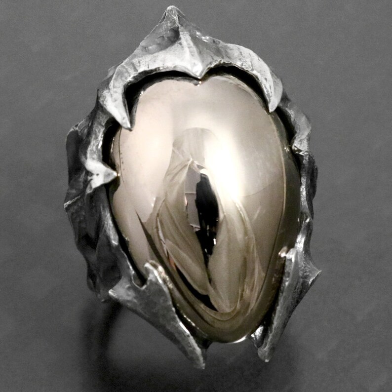 May include: A silver ring with a large, smooth, oval-shaped gemstone set in a textured, spiky, silver setting.