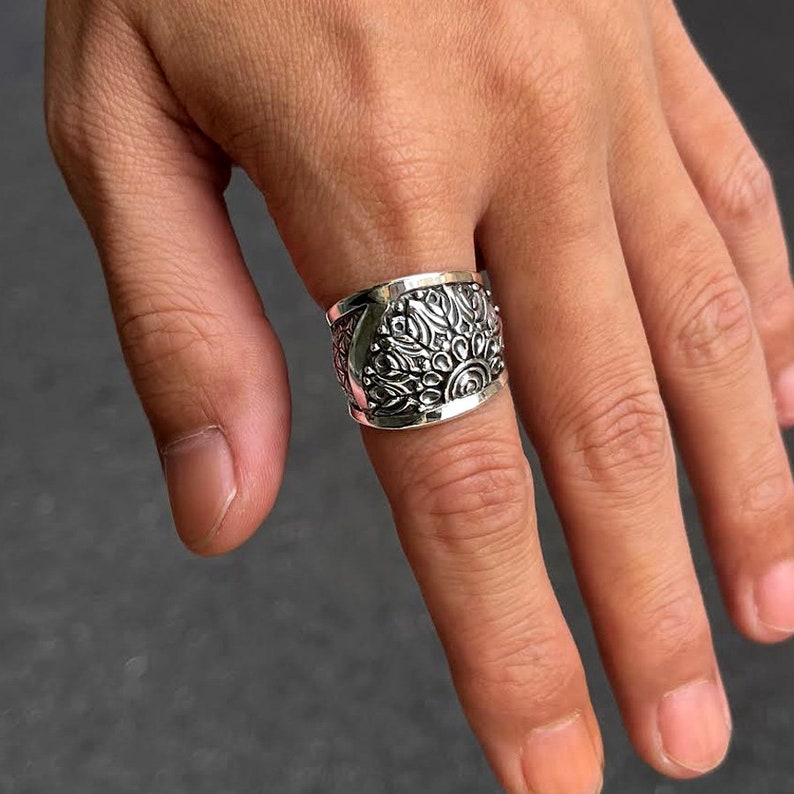May include: A silver ring with a wide band and an intricate floral design. The ring is worn on a finger.