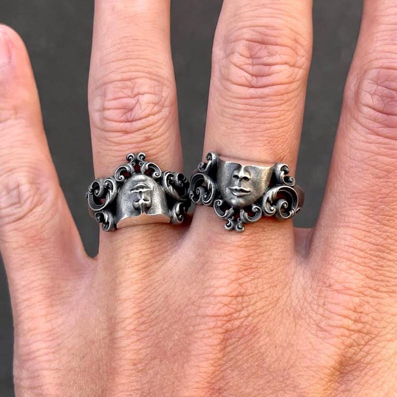 K&ouml;nnte beinhalten: Zwei silberne Ringe mit detaillierten Designs. Ein Ring zeigt ein stilisiertes Auge, der andere ein Gesicht. Beide Ringe haben verzierte, wirbelnde Muster um die zentralen Figuren, was eine Vintage- oder Gothic-&Auml;sthetik erzeugt.