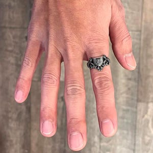 K&ouml;nnte beinhalten: Ein silberner Ring mit einem detaillierten Gesichtsdesign und kunstvollen Schn&ouml;rkeln. Der Ring wird an einem Finger getragen und unterstreicht seine aufwendige Handwerkskunst und seinen Vintage-Stil. Das Gesichtsdesign ist der Mittelpunkt, mit einer detaillierten Darstellung eines menschlichen Gesichts.