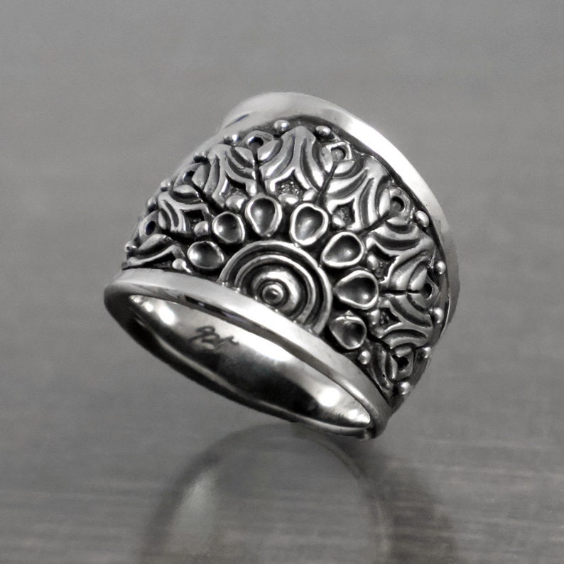 May include: A wide silver band ring with an intricate, ornate design featuring a circular pattern of raised dots and swirls.