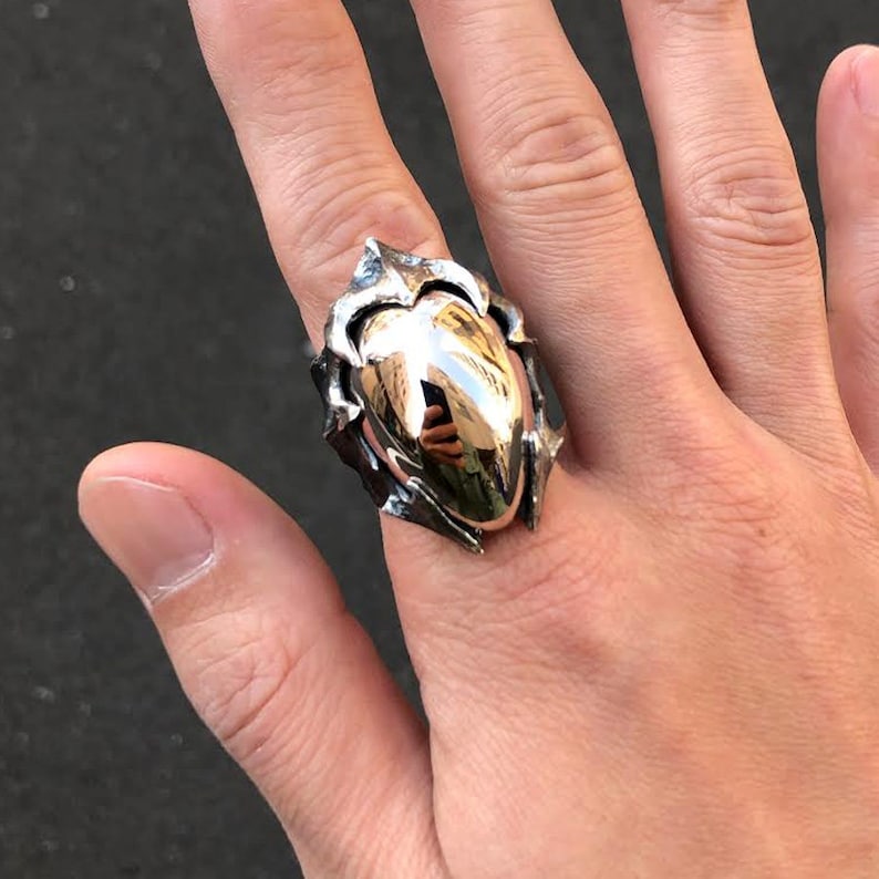 May include: A silver ring with a large, heart-shaped, polished, gold-toned center piece. The ring has a detailed, textured, and intricate design.