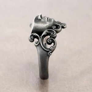 K&ouml;nnte beinhalten: Ein silberner Ring mit einem skulptierten Gesichtsprofil, das filigrane Details und dekorative Schn&ouml;rkel zeigt. Der Ring hat ein massives, sich verj&uuml;ngendes Band. Das Design erinnert an klassische Kunst, mit Fokus auf Gesicht und Verzierungen.