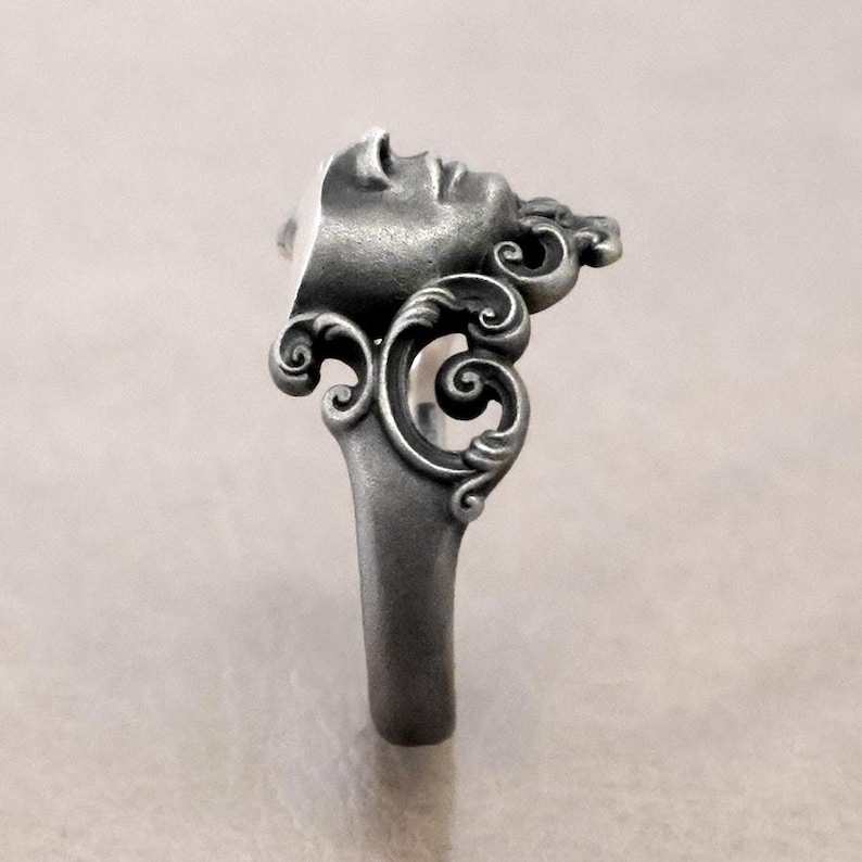 K&ouml;nnte beinhalten: Ein silberner Ring mit einem skulptierten Gesichtsprofil, das filigrane Details und dekorative Schn&ouml;rkel zeigt. Der Ring hat ein massives, sich verj&uuml;ngendes Band. Das Design erinnert an klassische Kunst, mit Fokus auf Gesicht und Verzierungen.