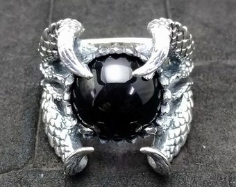 May include: A silver ring with a black gemstone set in a claw design. The claws are shaped like dragon claws and are detailed with scales.