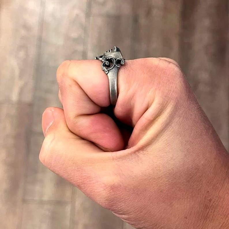 K&ouml;nnte beinhalten: Ein silberfarbener Ring mit einem verzierten Design, das ein geformtes Gesicht und wirbelnde Details aufweist. Der Ring wird an einer Hand getragen und zeigt seine aufwendige Handwerkskunst und seinen Vintage-Stil. Das Design des Rings ist detailliert und auff&auml;llig.