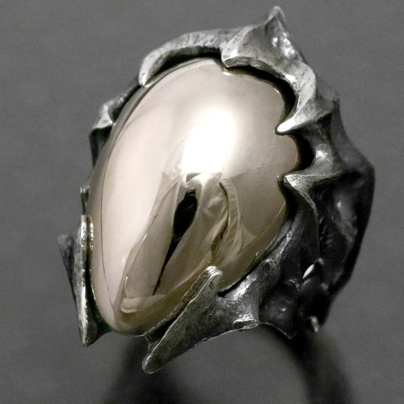 May include: A silver ring with a large, smooth, oval-shaped gemstone set in a textured, jagged, claw-like setting.