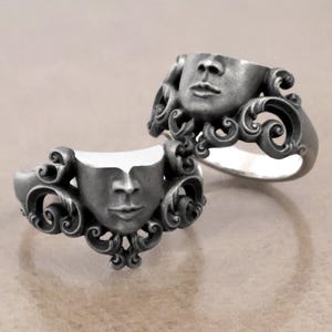 K&ouml;nnte beinhalten: Zwei silberfarbene Ringe mit einem Gesichtsdesign. Jeder Ring zeigt ein geformtes Gesicht mit detaillierten Gesichtsz&uuml;gen und verzierten, wirbelnden Verzierungen um das Gesicht. Die Ringe werden auf einem neutralen Hintergrund pr&auml;sentiert.