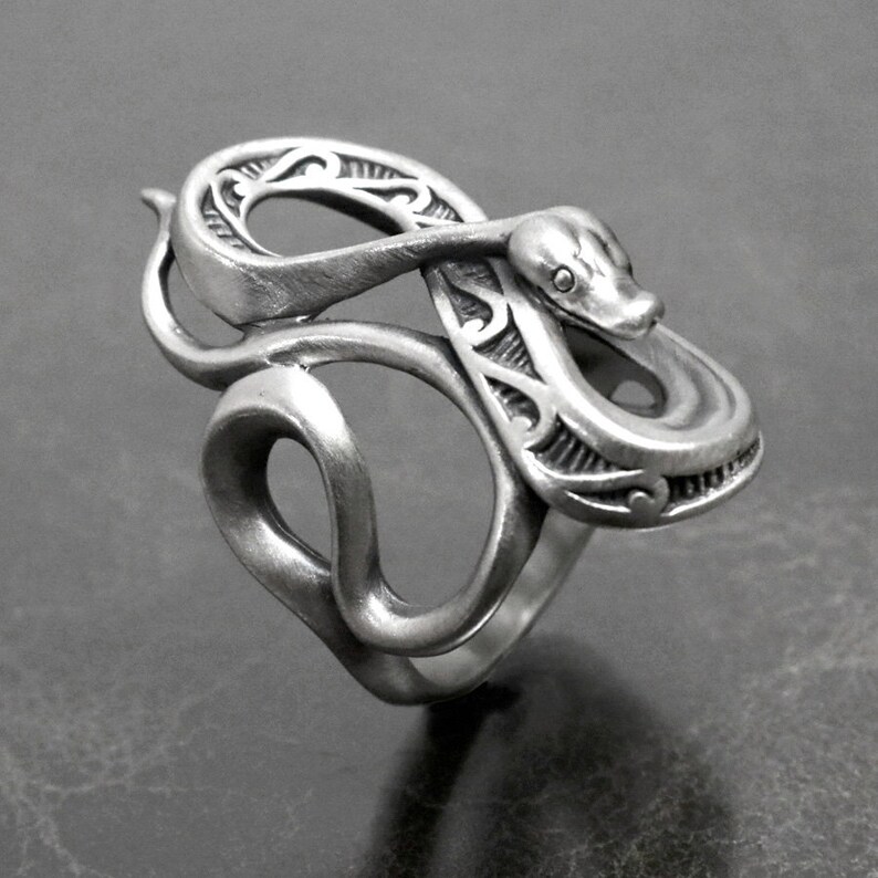 Op de afbeelding: Een zilveren slangenring met ingewikkelde details. De slang is opgerold en zijn kop is opgeheven, met een klein oogdetail. De ring is gemaakt van glad, gepolijst metaal.