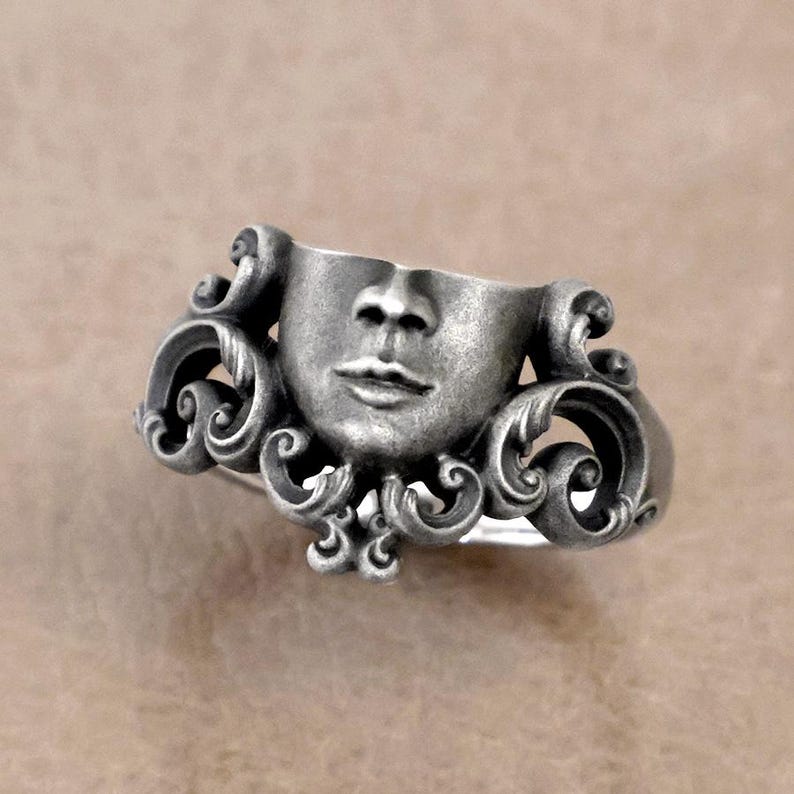 K&ouml;nnte beinhalten: Silberfarbener Ring mit einer modellierten Gesichtsform und detaillierten Gesichtsz&uuml;gen. Der Ring hat ein verziertes, wirbelndes Design um das Gesicht, was eine einzigartige, k&uuml;nstlerische &Auml;sthetik erzeugt. Der Ring ist wahrscheinlich aus Metall.