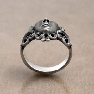 K&ouml;nnte beinhalten: Ein silberner Ring mit einem geformten Gesicht als zentralem Design. Der Ring hat detaillierte Gesichtsz&uuml;ge und dekorative, wirbelnde Muster. Ein einzigartiges Schmuckst&uuml;ck.