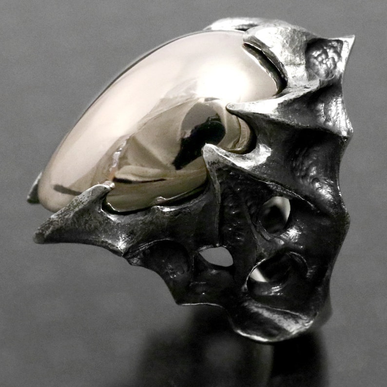 May include: A silver ring with a large, smooth, rounded, gold-colored stone set in a textured, jagged, claw-like setting.