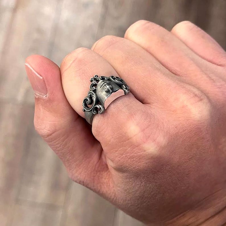 K&ouml;nnte beinhalten: Ein silberner Ring mit einem geformten Gesicht und aufwendigen Details. Der Ring hat eine Vintage-&Auml;sthetik mit einem detaillierten Gesichtsdesign und wirbelnden Mustern um den Kopf. Das Gesicht hat einen ruhigen Ausdruck und ist vor einem rosafarbenen Hintergrund platziert.