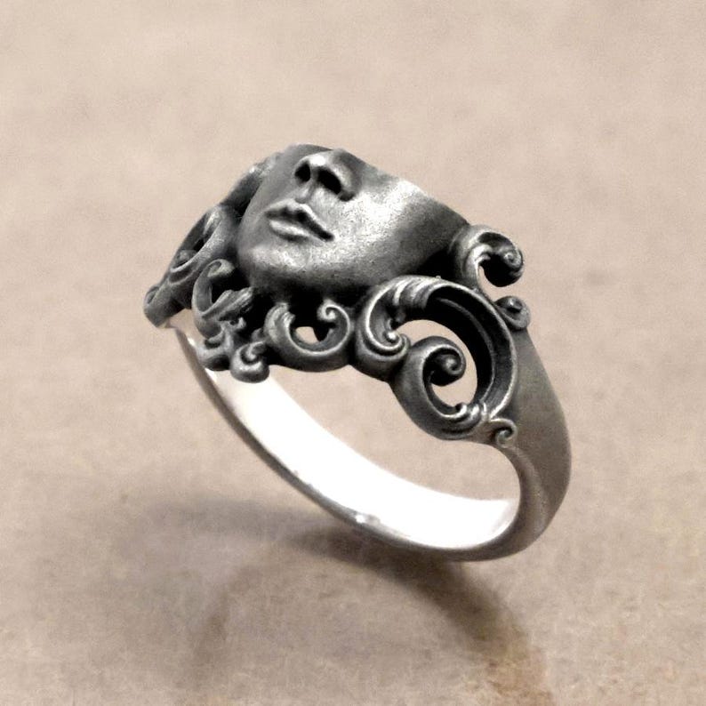 K&ouml;nnte beinhalten: Ein silberfarbener Ring mit einer modellierten Gesichtsform, der detaillierte Gesichtsz&uuml;ge und kunstvolle, wirbelnde Verzierungen aufweist. Das Design des Rings umfasst ein Teilgesicht und dekorative Verzierungen, wodurch ein einzigartiger Schmuck entsteht.