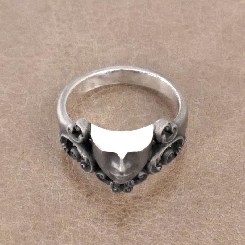 K&ouml;nnte beinhalten: Silberner Ring mit einem stilisierten Gesichtsdesign. Der Ring hat ein detailliertes Gesicht mit verzierten Schn&ouml;rkeln auf beiden Seiten. Der Ring besteht aus einem silberfarbenen Metall und hat eine polierte Oberfl&auml;che. Der Ring ist ein Schmuckst&uuml;ck.