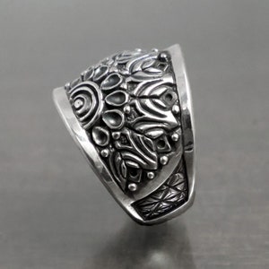 May include: A silver ring with a detailed, intricate design. The ring is wide and has a rounded top. The design features a variety of shapes and patterns, including circles, swirls, and leaves.