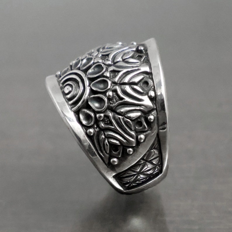May include: A silver ring with a detailed, intricate design. The ring is wide and has a rounded top. The design features a variety of shapes and patterns, including circles, swirls, and leaves.