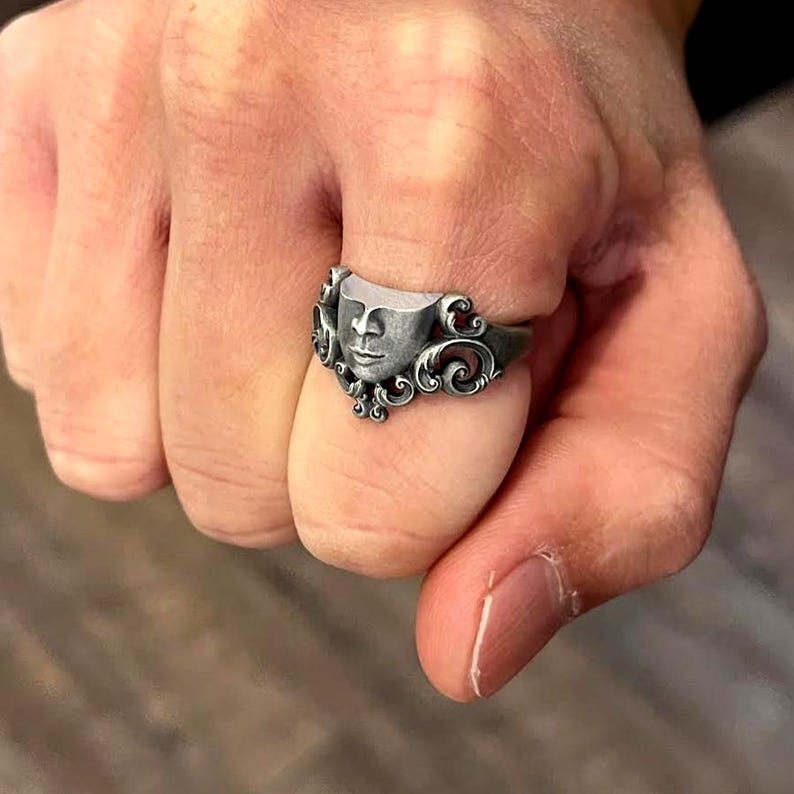 K&ouml;nnte beinhalten: Ein silberner Ring mit einem stilisierten Gesicht und aufwendigen Schn&ouml;rkeln. Der Ring wird an einem Finger getragen und zeigt das detaillierte Design und die Handwerkskunst des Schmucks.