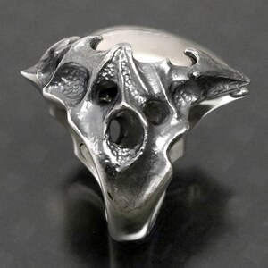 May include: A silver ring with a detailed, textured design resembling a skull or a creature's head. The ring has a large, open space in the center and a pointed, angular shape.