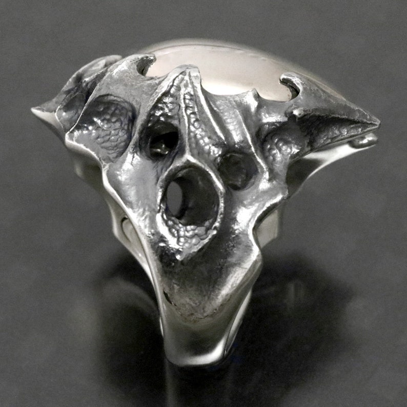 May include: A silver ring with a detailed, textured design resembling a skull or a creature's head. The ring has a large, open space in the center and a pointed, angular shape.