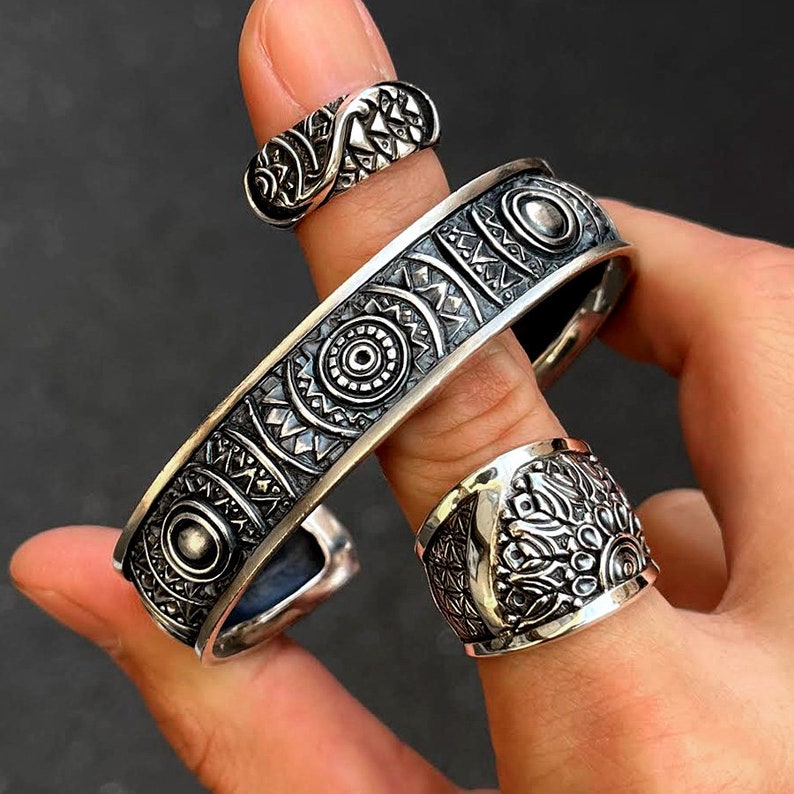 May include: Two silver rings with intricate geometric designs. The rings are worn on a finger and feature a circular pattern with raised details.