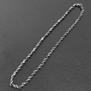 Puede incluir: Un collar de cadena de cuerda plateada. La cadena está hecha de eslabones de metal retorcidos, creando una apariencia de cuerda. El collar se muestra sobre un fondo gris oscuro.