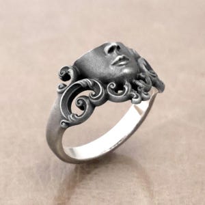 K&ouml;nnte beinhalten: Silberner Ring mit einer modellierten Gesichtsform, detaillierten Gesichtsz&uuml;gen und flie&szlig;endem Haar. Der Ring hat ein verziertes, wirbelndes Design um das Gesicht und einen glatten, polierten Ring. Ein einzigartiges Schmuckst&uuml;ck.