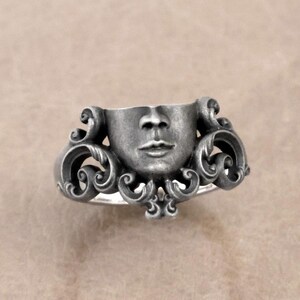 K&ouml;nnte beinhalten: Silberner Ring mit einer modellierten Gesichtsform und detaillierten Gesichtsz&uuml;gen. Der Ring hat ein verziertes, wirbelndes Design um das Gesicht herum, was ein einzigartiges Schmuckst&uuml;ck ergibt.