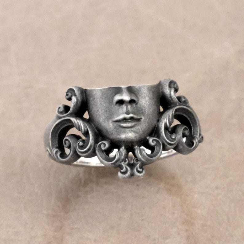 K&ouml;nnte beinhalten: Silberner Ring mit einer modellierten Gesichtsform und detaillierten Gesichtsz&uuml;gen. Der Ring hat ein verziertes, wirbelndes Design um das Gesicht herum, was ein einzigartiges Schmuckst&uuml;ck ergibt.