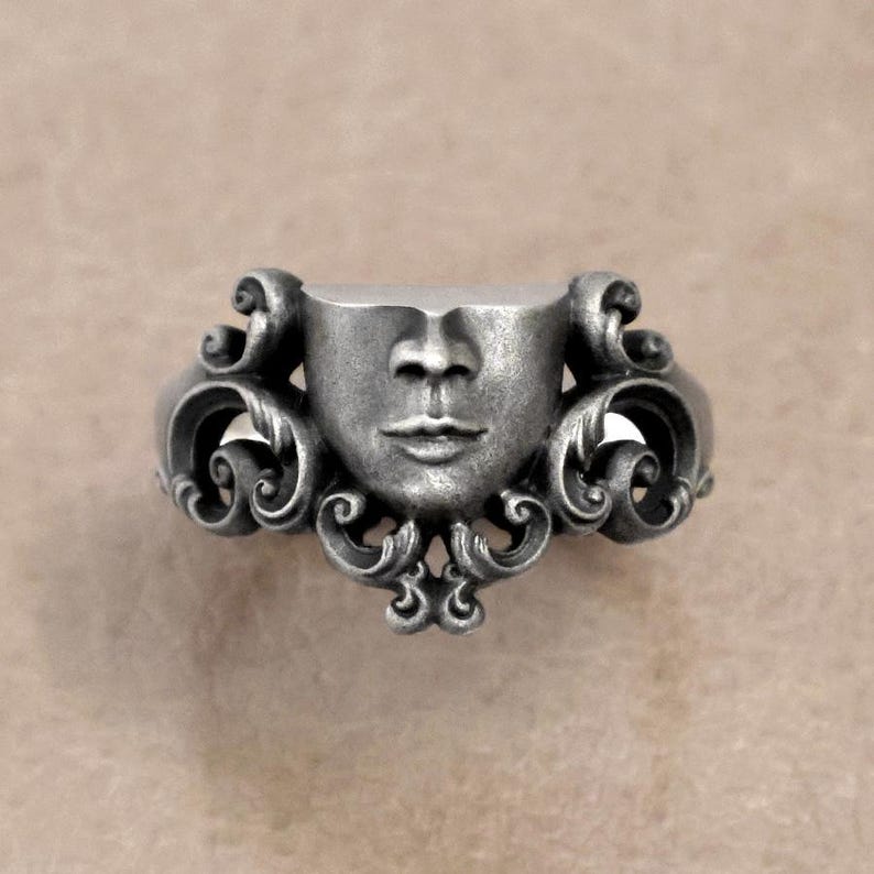 K&ouml;nnte beinhalten: Ein silberfarbener Ring mit einem geformten Gesicht mit detaillierten Gesichtsz&uuml;gen. Der Ring hat ein verziertes, wirbelndes Design auf beiden Seiten des Gesichts, das eine Vintage- oder Gothic-&Auml;sthetik verleiht. Der Ring ist wahrscheinlich aus Metall.