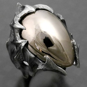 May include: A silver ring with a large, smooth, oval-shaped, gold-colored stone set in a textured, spiky, silver setting.