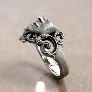 K&ouml;nnte beinhalten: Ein silberner Ring mit einem geformten Gesicht, das detaillierte Gesichtsz&uuml;ge und flie&szlig;endes Haar zeigt. Das Ringdesign enth&auml;lt verzierte, wirbelnde Muster um das Gesicht herum, was ihm eine klassische, k&uuml;nstlerische &Auml;sthetik verleiht. Der Ring besteht aus silberfarbenem Metall.