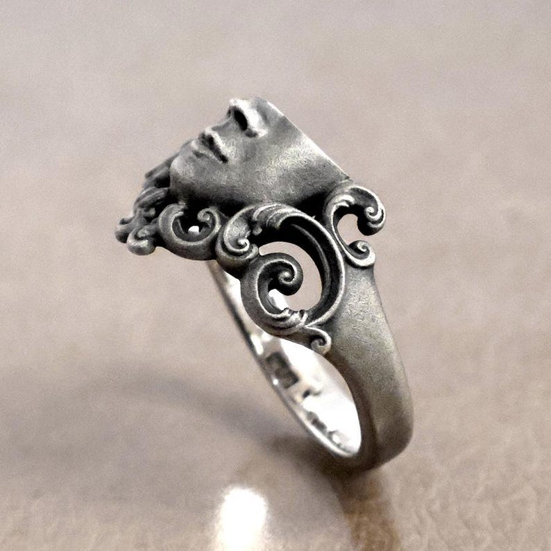 K&ouml;nnte beinhalten: Ein silberner Ring mit einem geformten Gesicht, das detaillierte Gesichtsz&uuml;ge und flie&szlig;endes Haar zeigt. Das Ringdesign enth&auml;lt verzierte, wirbelnde Muster um das Gesicht herum, was ihm eine klassische, k&uuml;nstlerische &Auml;sthetik verleiht. Der Ring besteht aus silberfarbenem Metall.