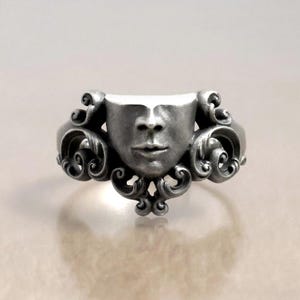 K&ouml;nnte beinhalten: Silberfarbener Ring mit einer modellierten Gesichtsform, oben teilweise verdeckt. Der Ring hat verzierte, wirbelnde Details an den Seiten und am Boden, die ein einzigartiges, k&uuml;nstlerisches Design schaffen. Das Gesicht des Rings ist ca. 2,5 cm breit.