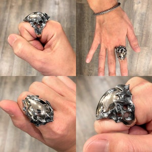 May include: A silver ring with a detailed, textured design resembling a skull or a creature. The ring is worn on a finger and is shown from multiple angles.