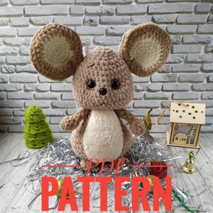 May include: Crochet pattern for a brown and white mouse amigurumi toy. The pattern is available as a PDF download. The image shows the finished toy against a brick wall background.