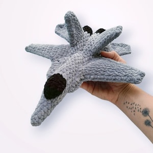 May include: A grey crocheted plush toy shaped like a fighter jet. The toy has black details on the nose and wings.