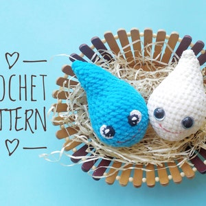 Può includere: Due gocce di pioggia all'uncinetto, una blu e una bianca, con facce sorridenti e occhietti tondi, sono annidate in un cesto intrecciato pieno di paglia.
