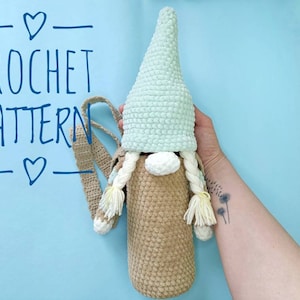 Könnte beinhalten: Häkelanleitung für einen Gnom-Flaschenhalter mit einem hellblauen Hut und einem beigefarbenen Körper. Der Gnom hat einen weißen Bart und gelbe Zöpfe. Der Flaschenhalter hat einen braunen Riemen.