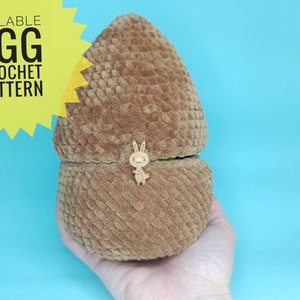 Könnte beinhalten: Ein braunes, befüllbares Häkeleimuster, das in einer Hand gehalten wird. Das Ei ist aus strukturiertem Garn gefertigt und hat einen Holzkaninchen-Knopfverschluss. Der Text "FILLABLE EGG CROCHET PATTERN" befindet sich in einem gelben Stern.