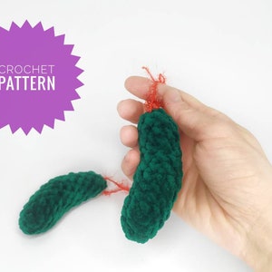 May include: Two green crocheted ornaments shaped like cucumbers with red yarn accents. The ornaments are held in a hand.