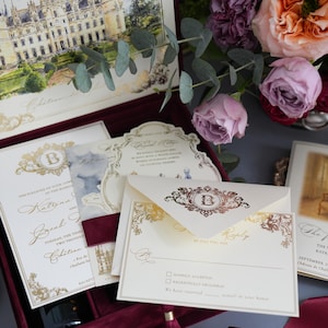 K&ouml;nnte beinhalten: Elegante Hochzeitseinladungs-Suite mit einer Aquarell-Schlossillustration, Goldfolienakzenten und einer bordeauxroten Samtbox. Enth&auml;lt Einladungskarten, Antwortkarten und Umschl&auml;ge. Blumenarrangement mit Rosen.