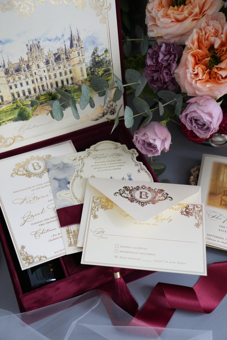 K&ouml;nnte beinhalten: Elegante Hochzeitseinladungs-Suite mit einer Aquarell-Schlossillustration, Goldfolienakzenten und einer bordeauxroten Samtbox. Enth&auml;lt Einladungskarten, Antwortkarten und Umschl&auml;ge. Blumenarrangement mit Rosen.