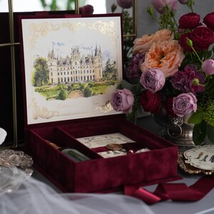 K&ouml;nnte beinhalten: Eine bordeauxfarbene Samt-Einladungsbox mit einer detaillierten Illustration eines Schlosses auf dem Deckel. Im Inneren sind Schreibwaren und ein Stift zu sehen. Ein Strau&szlig; Rosen und anderer Blumen steht in der N&auml;he, zusammen mit einer dekorativen B&uuml;ste und einem kleinen Tablett.
