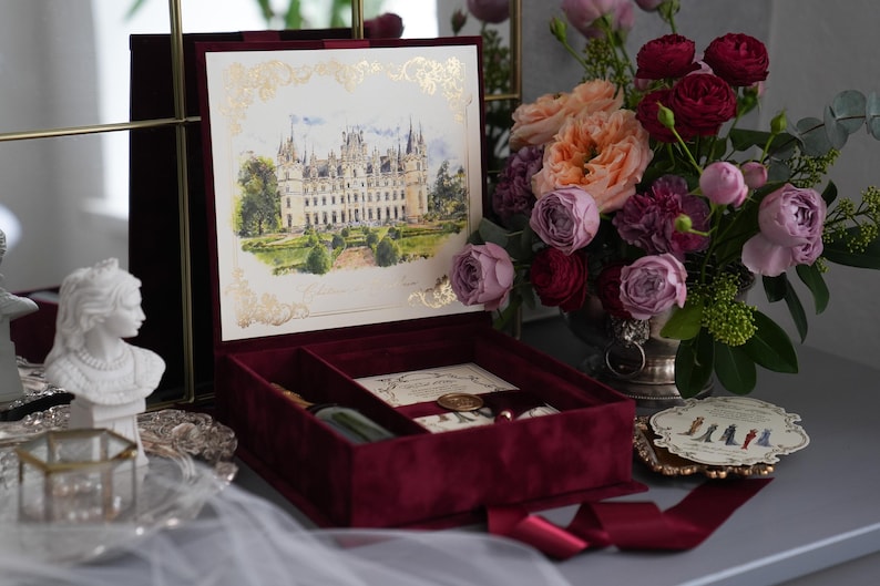 K&ouml;nnte beinhalten: Eine bordeauxfarbene Samt-Einladungsbox mit einer detaillierten Illustration eines Schlosses auf dem Deckel. Im Inneren sind Schreibwaren und ein Stift zu sehen. Ein Strau&szlig; Rosen und anderer Blumen steht in der N&auml;he, zusammen mit einer dekorativen B&uuml;ste und einem kleinen Tablett.