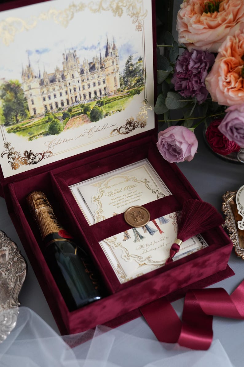 K&ouml;nnte beinhalten: Burgunderfarbene Samt-Pr&auml;sentationsbox mit Champagnerflasche, Einladungskarten und Quaste. Der Deckel der Box zeigt ein Aquarellgem&auml;lde eines Schlosses. Blumen im Hintergrund.