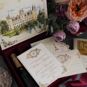K&ouml;nnte beinhalten: Hochzeitseinladungs-Set mit Schlossillustration, Goldakzenten und einer bordeauxroten Samtbox. Enth&auml;lt Einladungskarten, Umschl&auml;ge und eine Champagnerflasche. Blumen im Hintergrund.